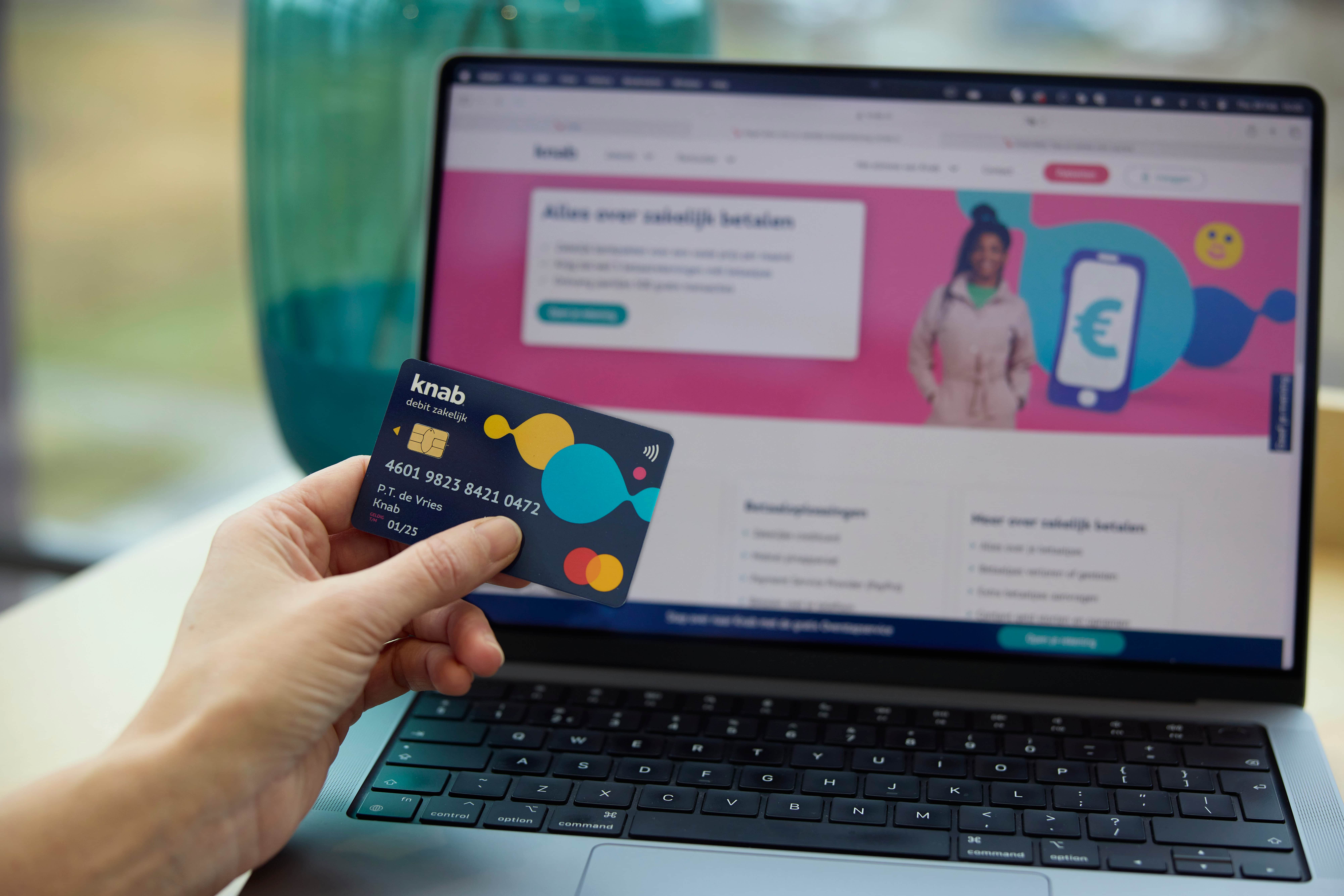 Hand houdt een Knab-debitkaart voor een laptop met de Knab bankwebsite op het sc